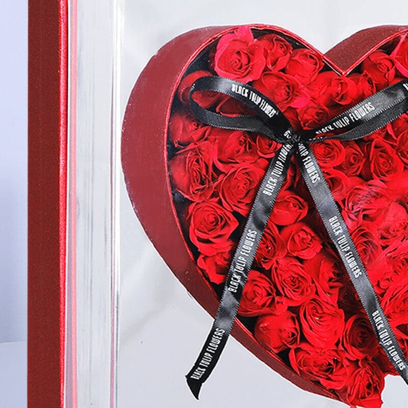 Close up view of heart-shaped red roses presented in a transparent box adorned with a black ribbon.