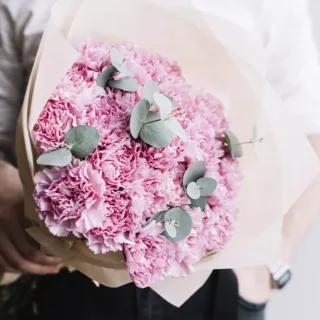 A bouquet of pink carnations with eucalyptus leaves, wrapped in beige paper.
