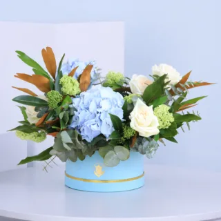 Elegant blue hydrangea, white roses, greenery, viburnum berries, and magnolia leaves in a chic blue round box.