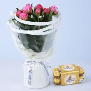 A bouquet of pink peonies, arranged in a simple, elegant style. The flowers are wrapped in white tissue paper and tied with a white ribbon. A box of Ferrero Rocher chocolates is placed beside the bouquet.
