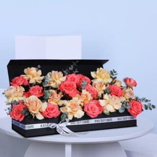Peach and coral roses nestled with eucalyptus, elegantly arranged in a rectangular black box and tied with a white ribbon.