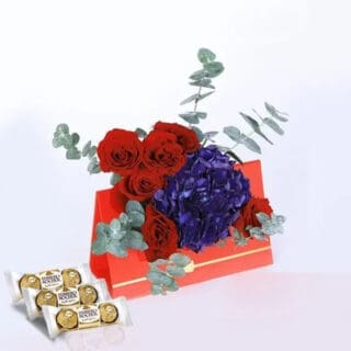 A red card with red roses, blue hydrangeas, and eucalyptus leaves, accompanied by a box of chocolates on the table below.