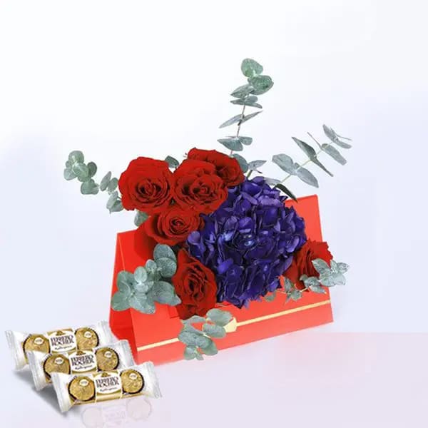01 (7) A red card with red roses, blue hydrangeas, and eucalyptus leaves, accompanied by a box of chocolates on the table below.