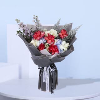 A stunning hand bouquet featuring roses, hydrangeas, delphinium, and seasonal fillers, wrapped in grey and tied with a black and white ribbon.