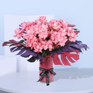 Arrangement with cluster of delicate pink hydrangeas surrounded by lush monstera leaves in a sleek pink vase.