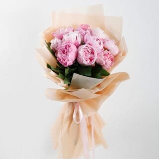 A hand-tied bouquet featuring lush, full-bloom pink peonies complemented by fresh green foliage with a brown wrap.