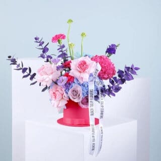 A mix of blue and pink hydrangeas, roses, lisianthus, carnations, ornithogalum, and chrysanthemum with eucalyptus in a pink hatbox tied with a white ribbon.