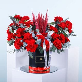 A stunning arrangement of red roses, red astilbe, and eucalyptus placed in a round black box adorned with a classic white ribbon.