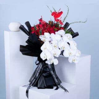 Red anthuriums, roses, red hypericums, white phalaenopsis orchids, and lush greenery in a black wrap tied with black ribbon.