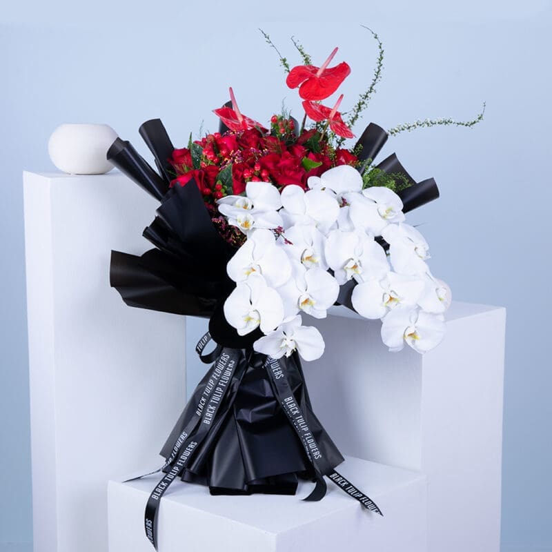 01 Red anthuriums, roses, red hypericums, white phalaenopsis orchids, and lush greenery in a black wrap tied with black ribbon.