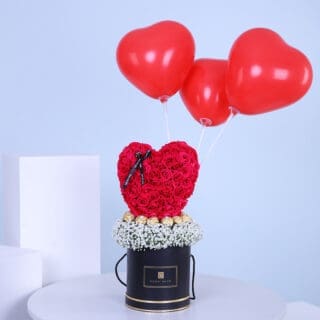 A black box with red roses in a heart shape, surrounded by white baby's breath, topped with three red heart-shaped balloons.