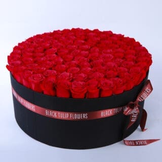 A sleek black circular box adorned with a red ribbon, filled with a stunning arrangement of vibrant red roses.