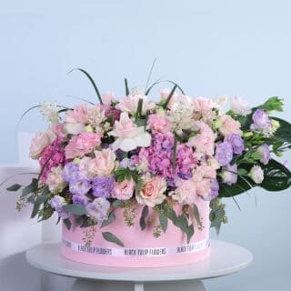 A large round arrangement of pink and purple roses, hydrangeas, lisianthus, and greenery in a pink box, tied with a white ribbon.