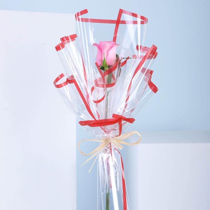 01 A single pink rose in a clear gift wrapping with red ribbons against a blue and white background.