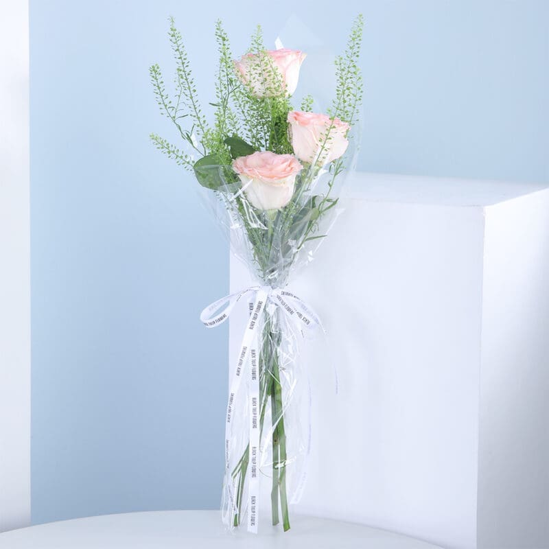 A bouquet of pink roses wrapped in cellophane with a white ribbon, on a white table against a blue background.