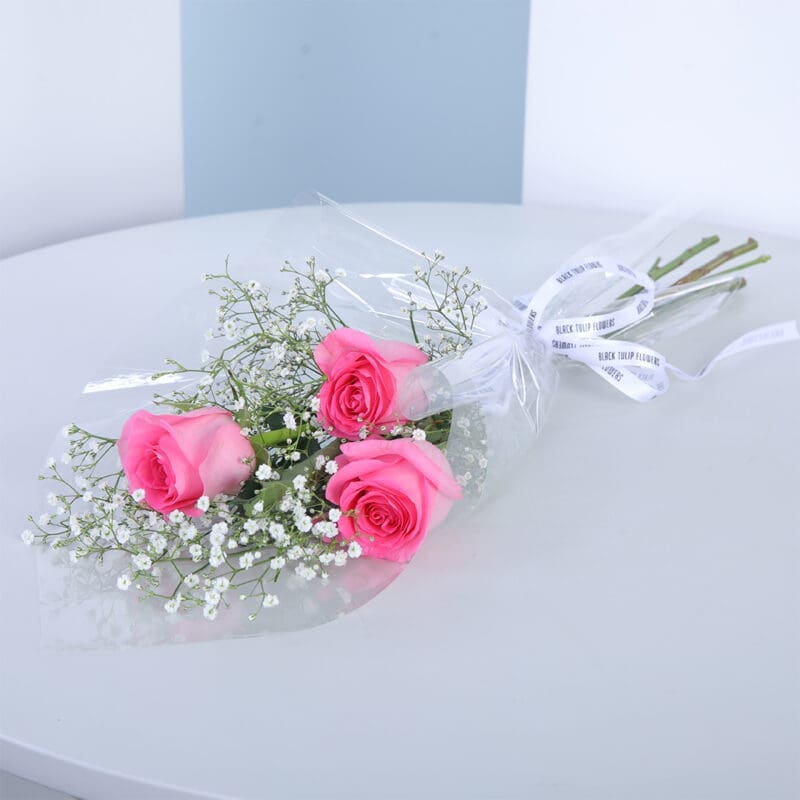 04 Small bouquet of 3 pink roses with baby's breath and greenery in a transparent wrap tied with white ribbon.