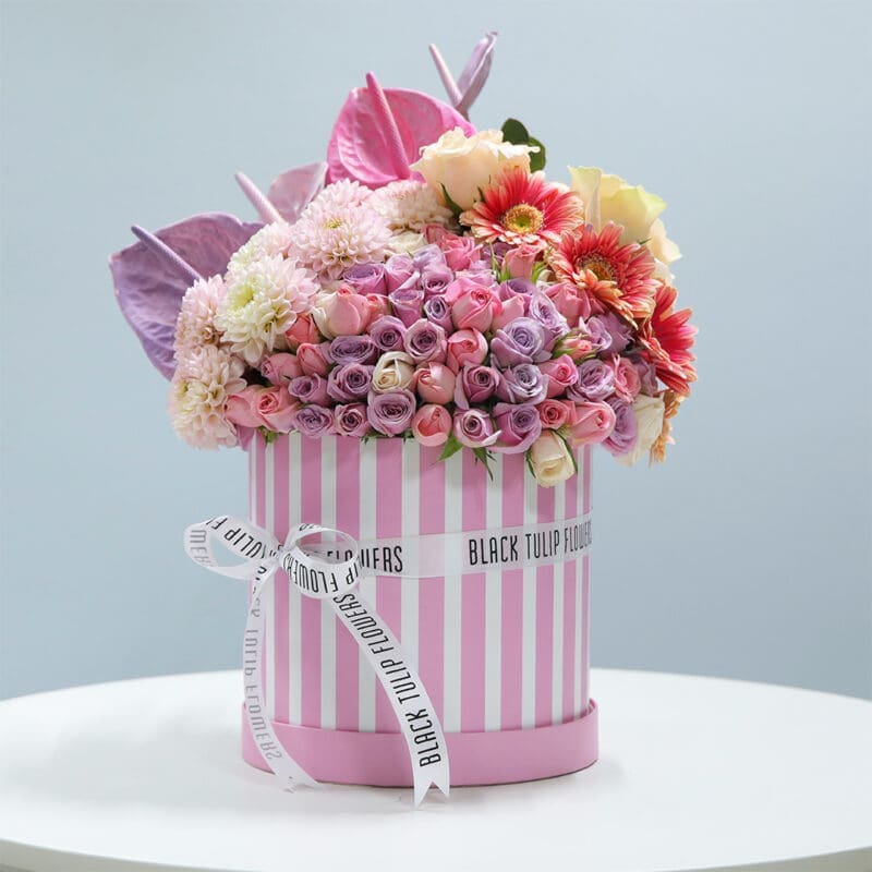 01 A pink striped cylindrical box filled with pink roses, purple spray roses, white chrysanthemums, pink anthuriums, and orange gerberas.