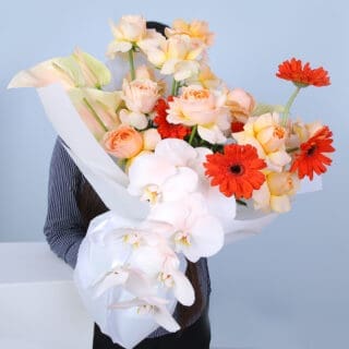 A vibrant bouquet featuring peach roses, white orchids, and red gerberas, wrapped in white paper