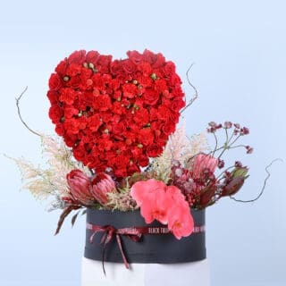 A round black box filled with a heart-shaped arrangement of 30 red roses, bunches of red carnations, protea stems, and more.