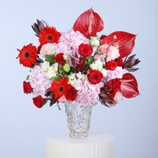 A floral arrangement in glass vase featuring red gerberas, pink hydrangeas, red roses, and striking anthurium leaves.