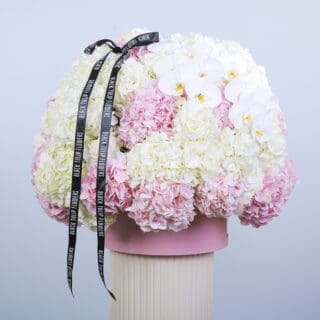 A hat box, featuring pink and white hydrangeas and white orchids with a black ribbon" black tulip flowers" on it