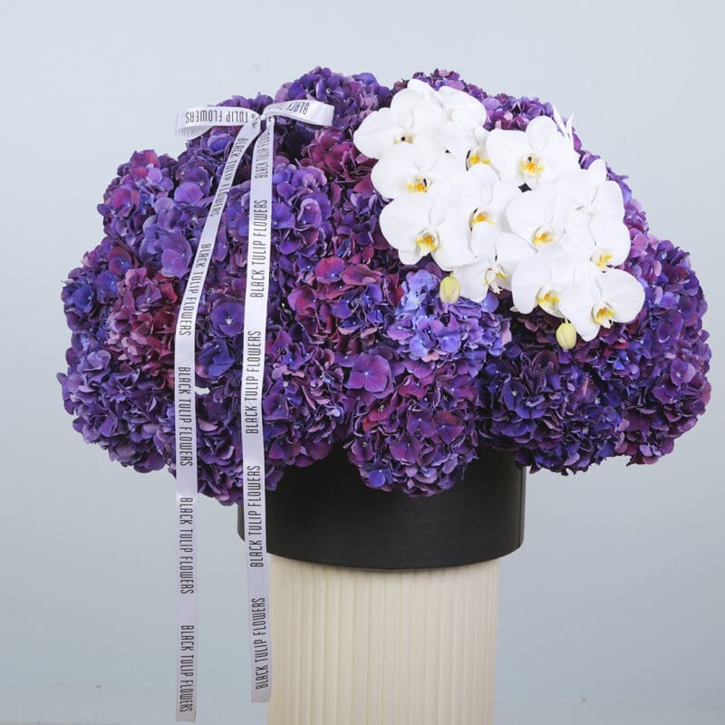 Floral arrangement in a hat box, featuring purple hydrangeas and white orchids.