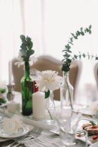 A beautifully set table with a minimalist, elegant aesthetic. White peonies and eucalyptus sprigs are arranged in recycled glass bottles as centerpieces. The table is set with white plates, silverware, and wine glasses. There are place cards with names written on them, adding a personal touch.