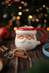 A festive Christmas mug shaped like Santa Claus's face, filled with a warm beverage. The mug is surrounded by cinnamon sticks, apples, and a blurred background of a Christmas tree with twinkling lights.