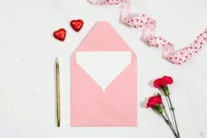 A pink envelope with a blank card inside, next to a pen, heart-shaped chocolates, and carnations.