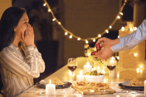 10 Gift Ideas to Kick Off the Valentine's Week in Style 11 At a dinner table, a man offers a ring to a woman, symbolizing a heartfelt proposal in an intimate setting