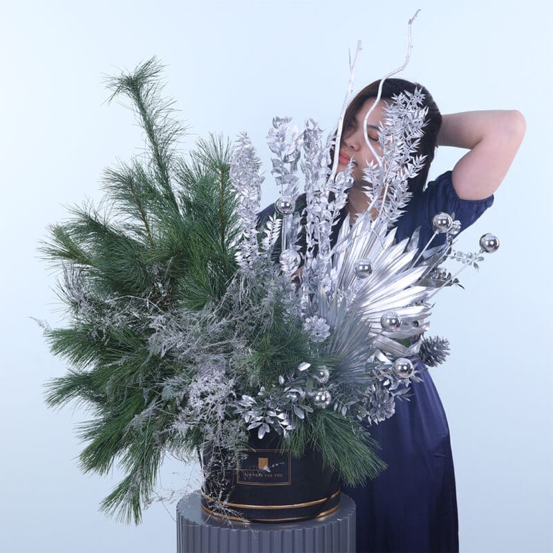 Silver-Glow-Holiday-Box A girl standing behind a box of green ferns and silver-coated pines for Christmas décor