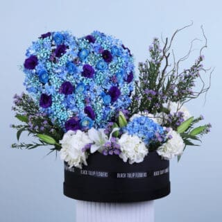 A Flower box featuring white and blue hydrangeas, roses, lisianthus, and aster arranged in a heart shape in a round black box.