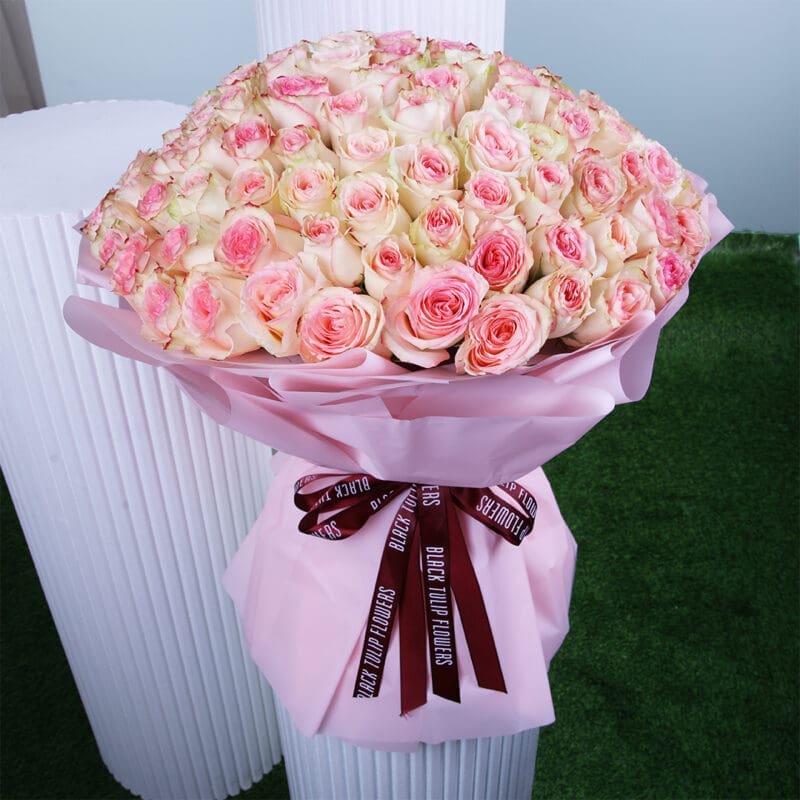 02 A bouquet of pink and white roses, wrapped in pink paper with a burgundy "BLACK TULIP FLOWERS" ribbon, photographed against a white column and green grass-like surface.