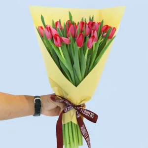 A bouquet of vibrant red tulips wrapped in yellow paper, held by a person's hand