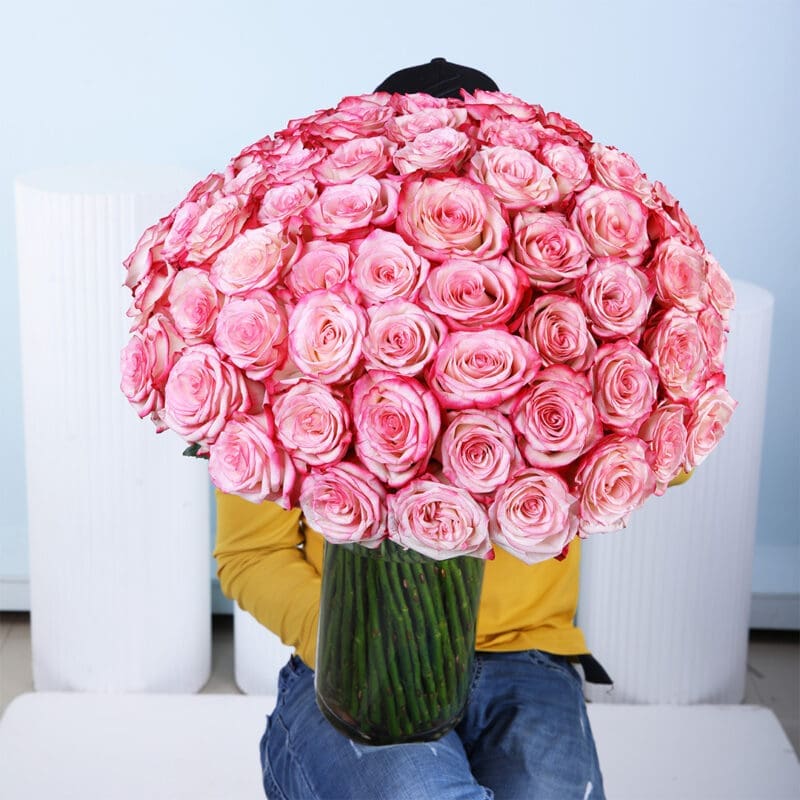 bi-colour-pink-rose-vase-main A large bouquet of 100 vibrant two-tone pink roses, with petals from a lighter pink at the center to a deeper, more saturated pink at the edges, is presented in a tall, clear glass cylindrical vase.