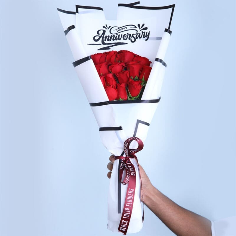 02 Bouquet of red roses wrapped in a white "Happy Anniversary" wrapper with a maroon ribbon, displayed on a white pedestal.