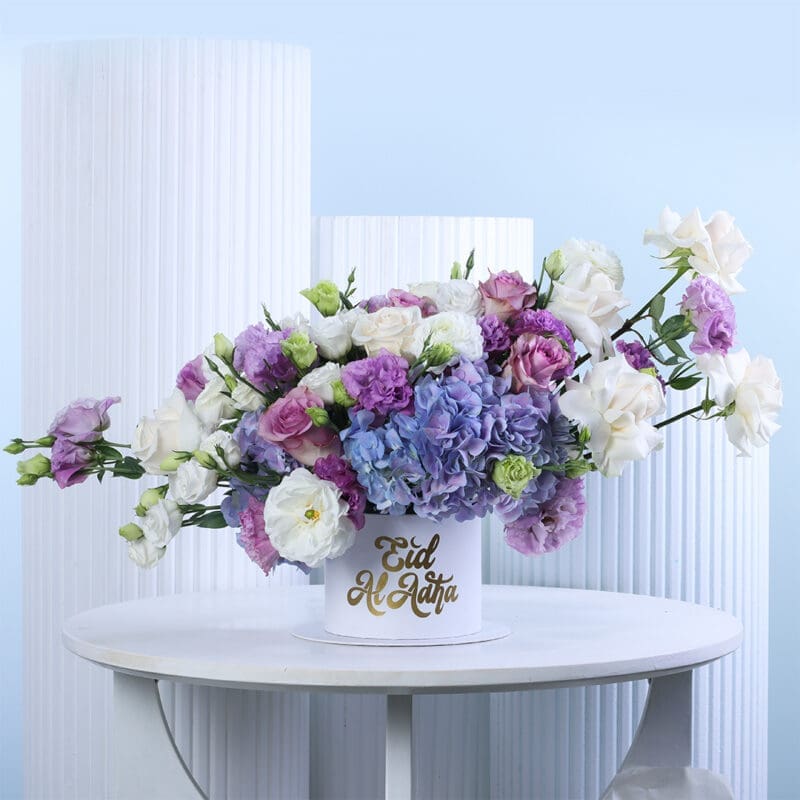 eid-luxury-box A luxurious floral arrangement featuring an assortment of purple and blue hydrangeas, white and light purple roses, and mixed blooms, presented in a white round box with "Eid Al Adha" written in gold script.