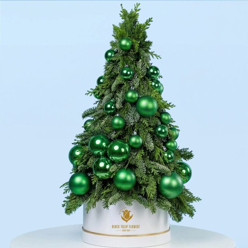 emerald-tabletop-christmas-tree Mini tabletop Christmas tree with lush green branches and shiny green baubles in a white pot on a round table.