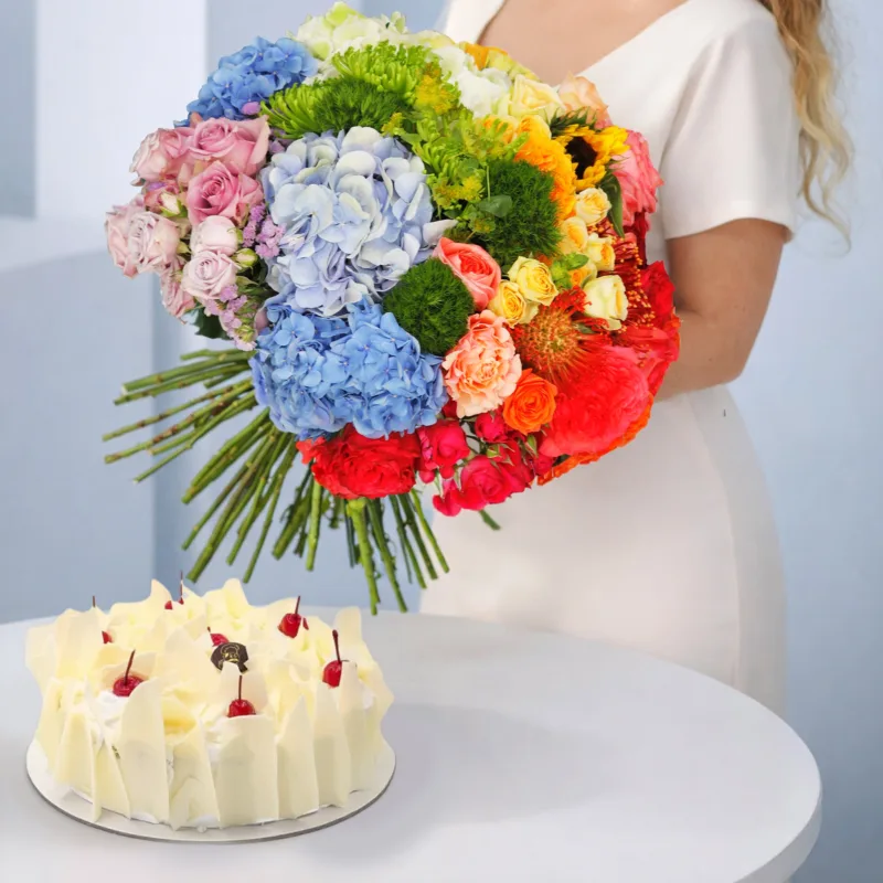 A gift box arrangement featuring a vibrant mixed flower bouquet with assorted fresh blooms placed neatly beside a white forest cake.