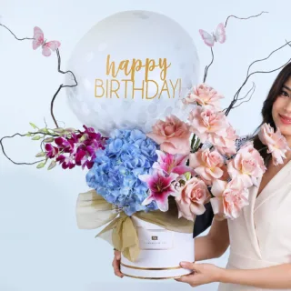 White round box decorated with blue hydrangea, purple orchid, cappuccino roses, lilium, ornamental butterflies, salix branches with a Happy Birthday balloon as a birthday surprise for a beloved one.