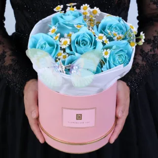 A decorative floral arrangement featuring blue roses, delicate white blooms, and a butterfly accent, elegantly arranged in a pink round box.
