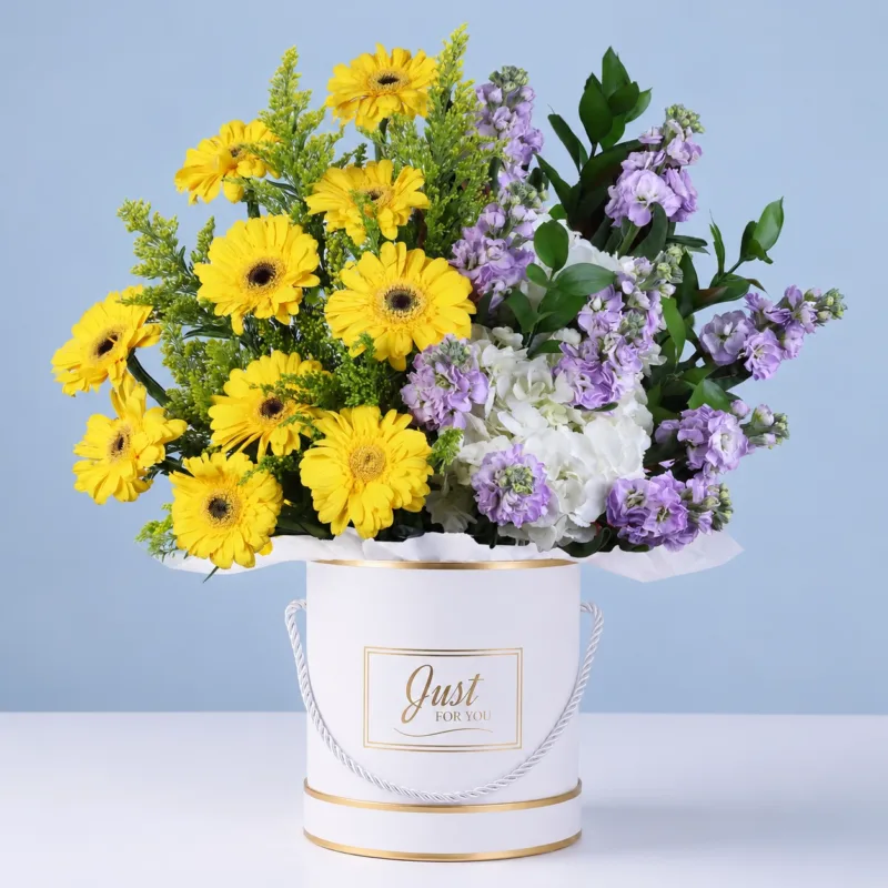 delightful-bloom-box Round box flower arrangement with cheerful yellow gerberas, soft lavender blooms, white hydrangeas, and fresh ruscus leaves for a premium floral gift.
