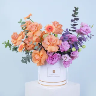 Blooms For You Hat Box featuring fresh peach roses, peach gerberas, purple lisianthus, and eucalyptus beautifully arranged in a premium hat box.