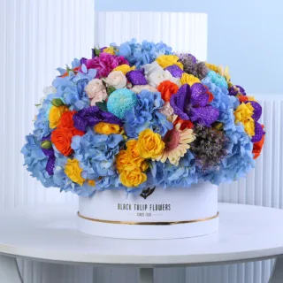 Blue Symphony Floral Hat Box featuring fresh blue hydrangeas, yellow spray roses, blue painted ping pong chrysanthemums, peach gerberas, purple mokara orchids, and mini eryngium beautifully arranged in a premium white hat box for gifting and special occasions.