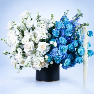 Blue & White Luxury Hat Box featuring fresh blue painted roses, White O'Hara roses, white matthiola, blue and white hydrangeas, blue delphiniums, white lisianthus, and dried amaranthus beautifully arranged in a premium black hat box for gifting and special occasions.