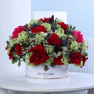 Crimson Elegance Floral Hat Box featuring fresh red roses, dark pink roses, viburnum, blue eryngium, white wax flowers, and pittosporum beautifully arranged in a premium white hat box for gifting and special occasions.