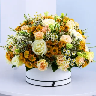 Golden Bloom Floral Hat Box featuring fresh orange santini chrysanthemums, white roses, peach lisianthus, brunia, white tanacetum, and white wax flowers beautifully arranged in a premium white hat box for gifting and special occasions.