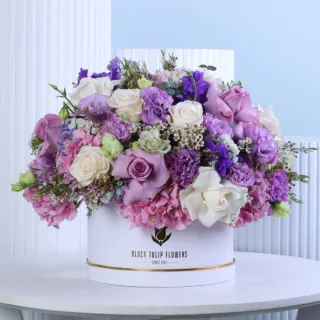 Lavender Blush Floral Hat Box featuring fresh cream roses, purple roses, pink hydrangeas, purple lisianthus, purple delphiniums, and white wax flowers beautifully arranged in a premium white hat box for gifting and special occasions.