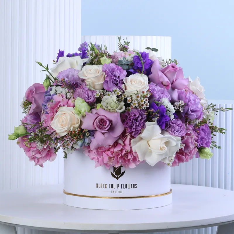 lavender-blush-floral-hat-box Lavender Blush Floral Hat Box featuring fresh cream roses, purple roses, pink hydrangeas, purple lisianthus, purple delphiniums, and white wax flowers beautifully arranged in a premium white hat box for gifting and special occasions.