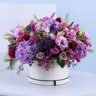 Lavender Luxe Floral Hat Box featuring fresh purple hydrangeas, purple trachelium, purple lisianthus, ping pong chrysanthemums, astilbe, statice, and wax flowers beautifully arranged in a premium white hat box for gifting and special occasions.